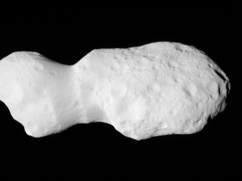 A close-up image of a white, irregularly shaped asteroid with a lumpy, cratered surface, set against the blackness of space.