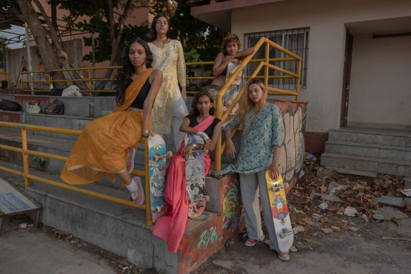 Five young women in colorful saris pose with skateboards by yellow railings and concrete steps in an urban outdoor setting, with trees and a building in the background.