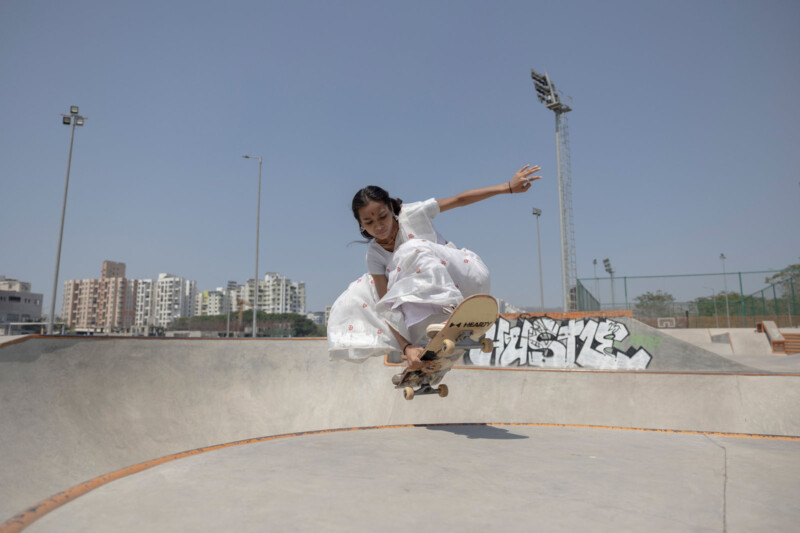 A young girl wearing a white dress performs a skateboard trick in a skate park, mid-air above a ramp, with buildings and graffiti visible in the background under a clear sky.