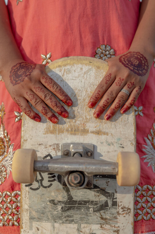 Close-up of two hands decorated with henna resting on a worn skateboard, with a coral-colored embroidered dress visible in the background.