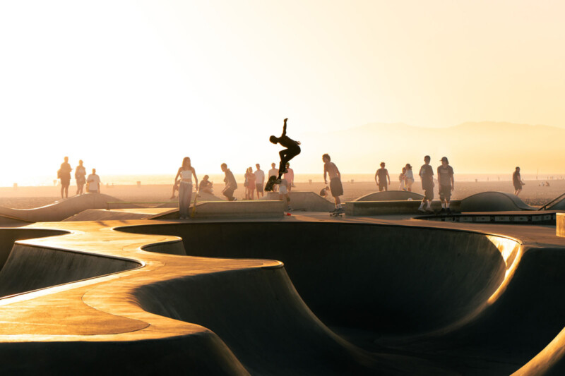 A skateboarder performs a trick in midair at a skate park during sunset, with several people walking and watching in the background under a hazy golden sky.