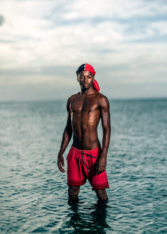 A man wearing red shorts and a red and black headscarf stands waist-deep in calm ocean water, with a long beaded necklace. The sky is cloudy and the horizon is visible in the background.