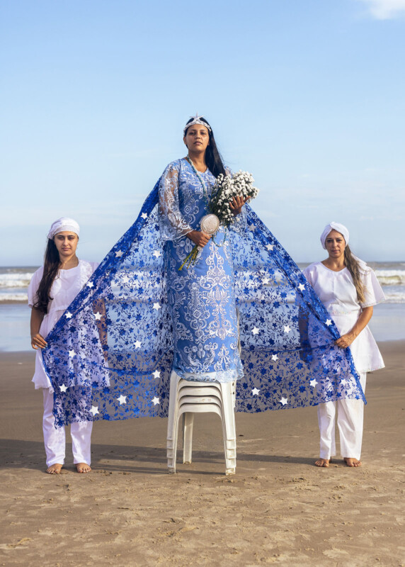 Three women stand on a beach. The central woman, elevated on stacked white chairs, wears a blue, star-patterned dress with a long cape, holding white flowers. Two women in white outfits hold the cape’s edges.