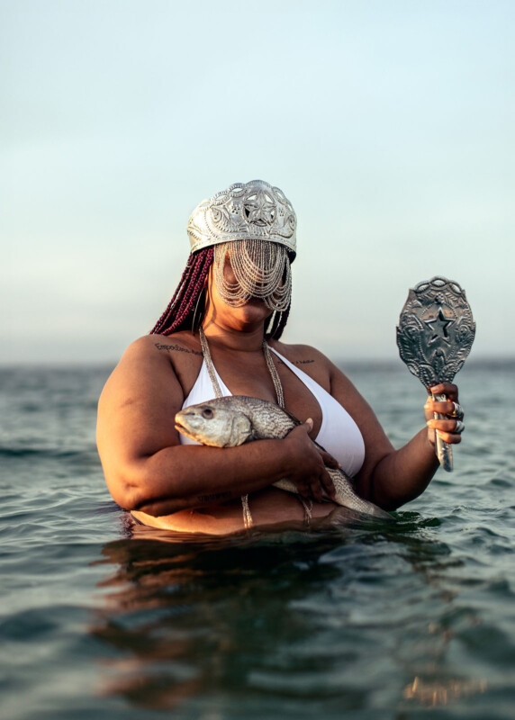 A person in a white bikini stands waist-deep in water, holding a silver fish in one arm and an ornate handheld mirror in the other. They wear a beaded headdress with chains covering their face.