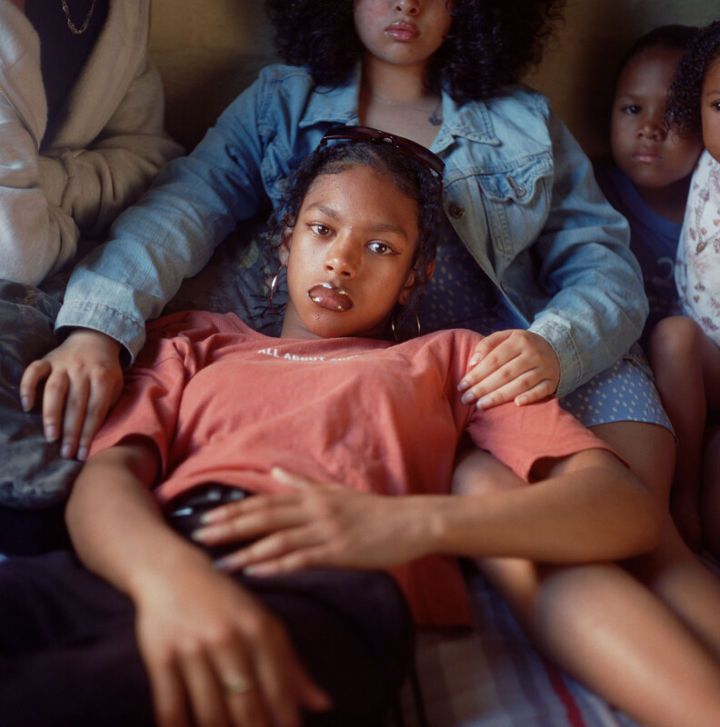 A young woman in an orange shirt lies on a bed, resting her head on another person's lap. Two other people sit close by, partially visible, creating a sense of comfort and togetherness.
