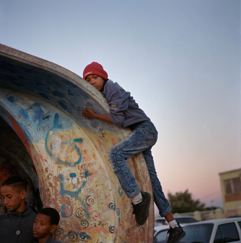 A boy wearing a red beanie and gray jacket climbs the side of a painted concrete structure, while two other boys stand below. The sky is clear and buildings are visible in the background.