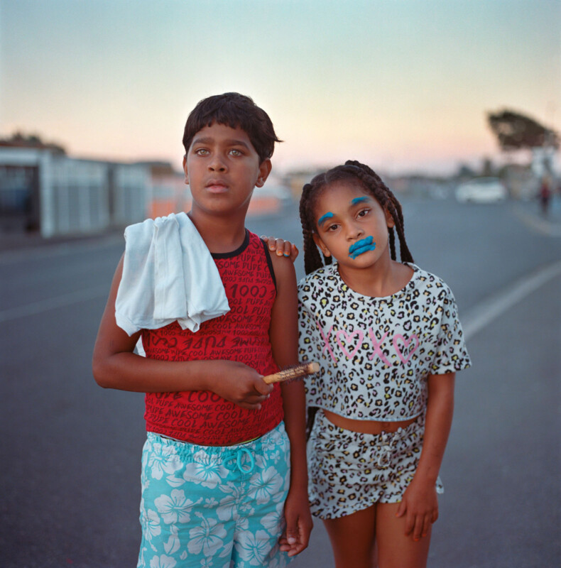 Two children stand together on a street at sunset. The boy wears a red shirt, blue floral shorts, and has a towel over his shoulder. The girl wears a leopard print outfit and bright blue lipstick. Both look directly at the camera.