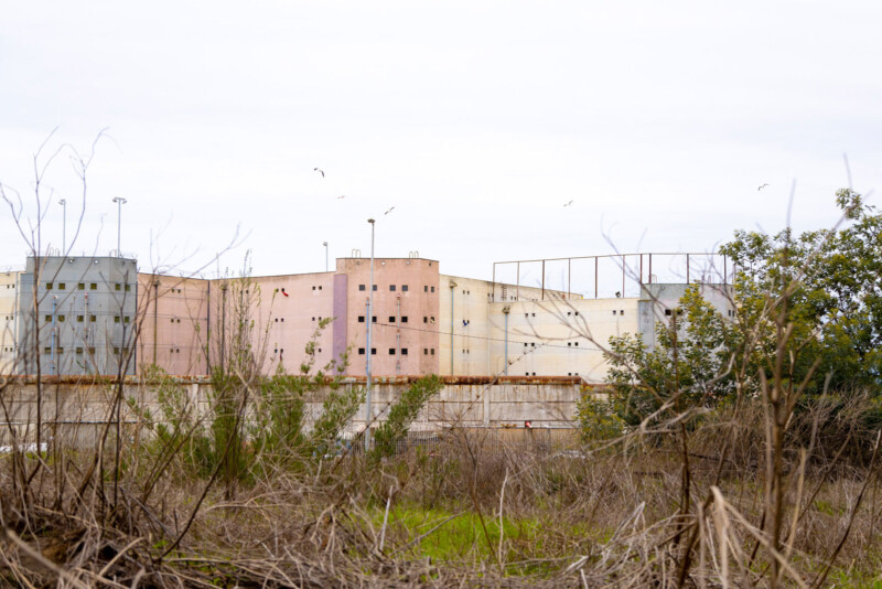 A distant view of a large, worn, pastel-colored building with barred windows, surrounded by a high fence, overgrown vegetation, and bare trees under a cloudy sky.