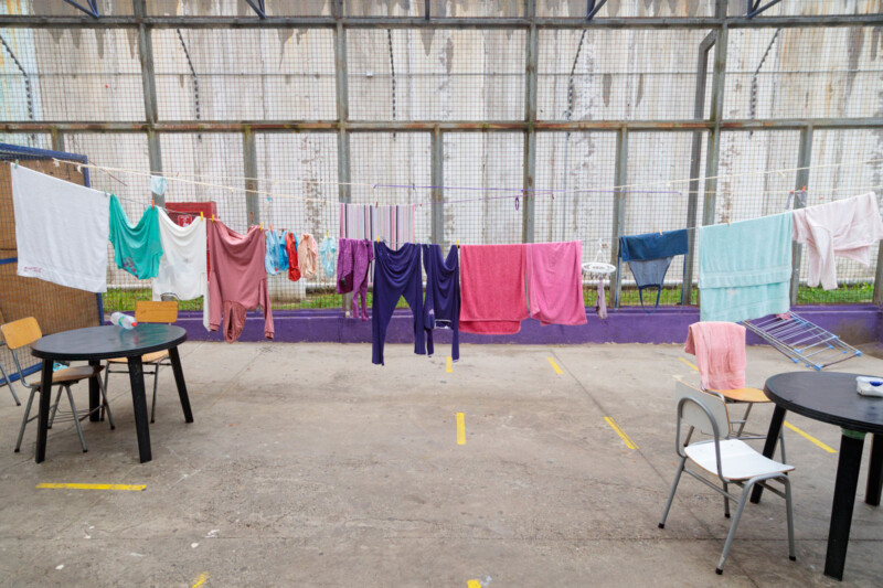 Clothes and towels in various colors hang on clotheslines in an outdoor concrete area with tables and chairs; a tall wire fence and a wall are in the background.