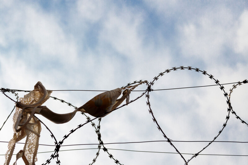 A bra and lace fabric hang tangled on barbed wire against a backdrop of a cloudy blue sky.