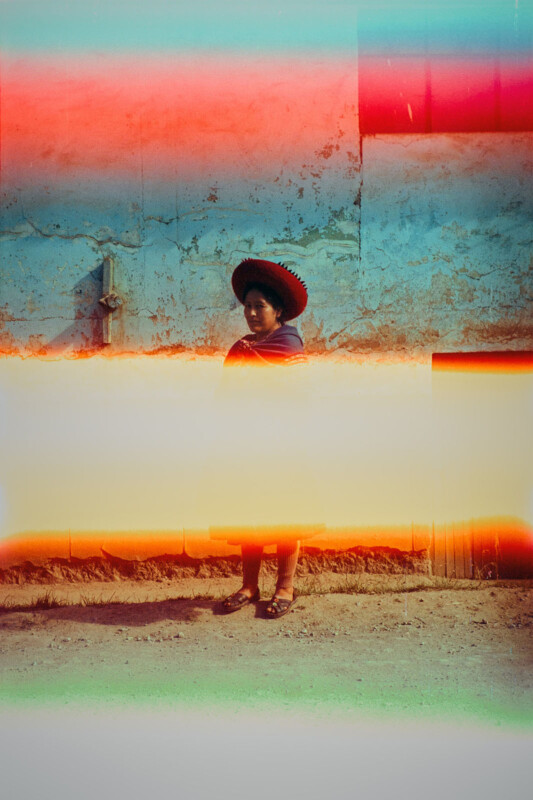 A person in traditional clothing and a red hat stands against a weathered blue wall. The image is partially obscured by colorful light leaks and film burns, creating bright horizontal bands across the photo.