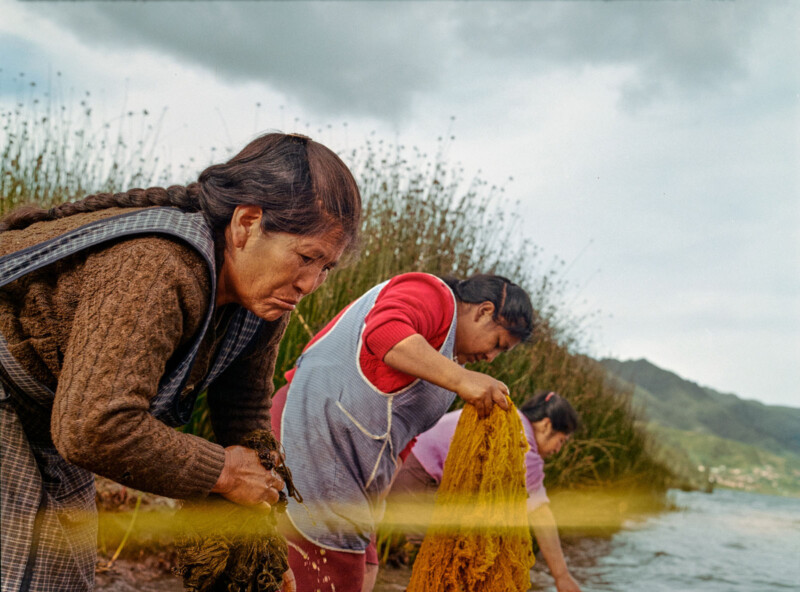 Three women stand at the edge of a lake, washing clothes by hand. They appear focused on their tasks, with tall grass and distant hills in the background under a cloudy sky.