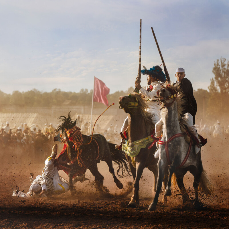 Three men in traditional clothing ride decorated horses at high speed on a dirt field, kicking up dust, while one rider falls to the ground. A red flag and a crowd are visible in the background.