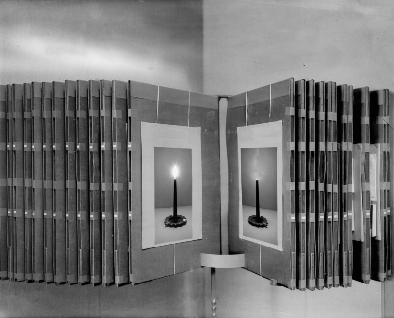 A display of open books arranged in a semicircle, each featuring an image of a single lit candle on its page, with a plain background and soft lighting.