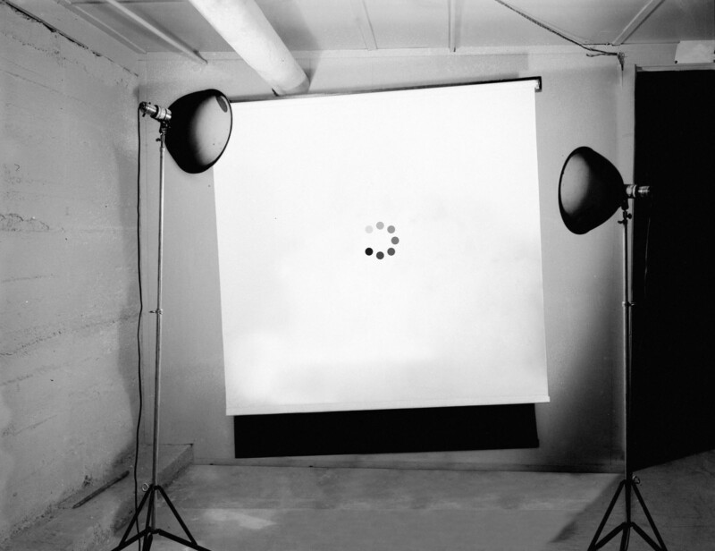 A black-and-white photo of a blank projection screen with a loading icon in the center, flanked by two studio lights on stands, set up in a bare room with concrete walls and floor.