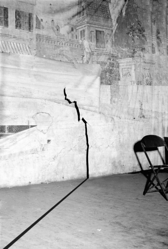 A large crack runs vertically up a worn, painted wall with a mural of buildings. Part of the mural is torn and peeling. Two empty folding chairs sit to the right on a bare floor.