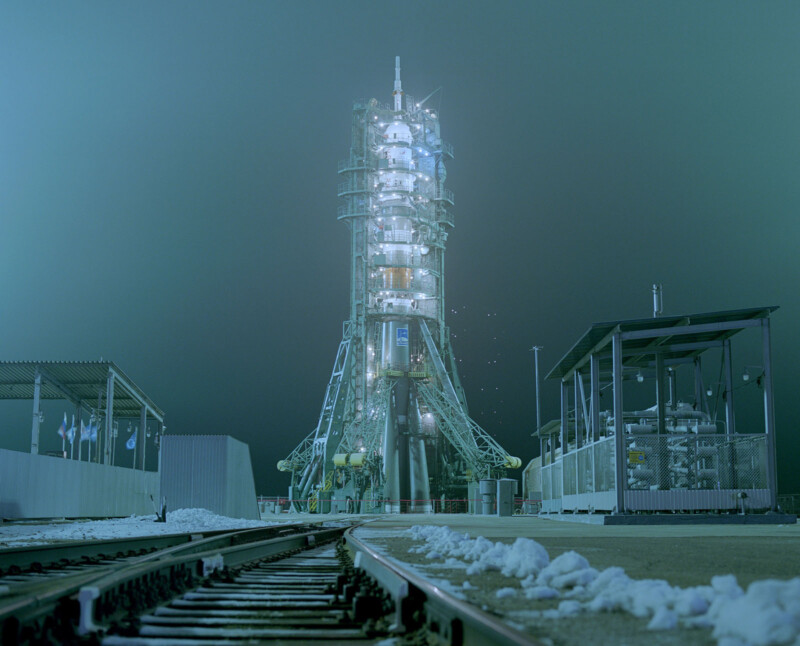 A brightly illuminated rocket stands on a launch pad at night, surrounded by metallic structures and snow-covered ground, with train tracks leading toward it in the foreground.