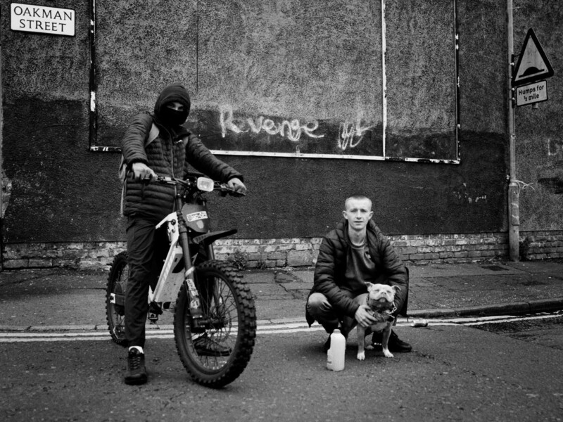 Two men pose on a street corner; one sits on a dirt bike wearing a mask, while the other crouches next to a dog. They are in front of a wall with graffiti reading "Revenge." Street signs are visible.