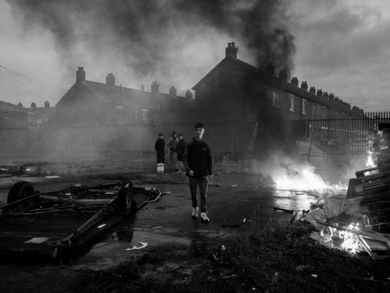 A young man walks through a smoky, debris-strewn urban lot at dusk, with fires burning and thick smoke rising. Behind him, several people stand together near terraced houses under a dark, cloudy sky.