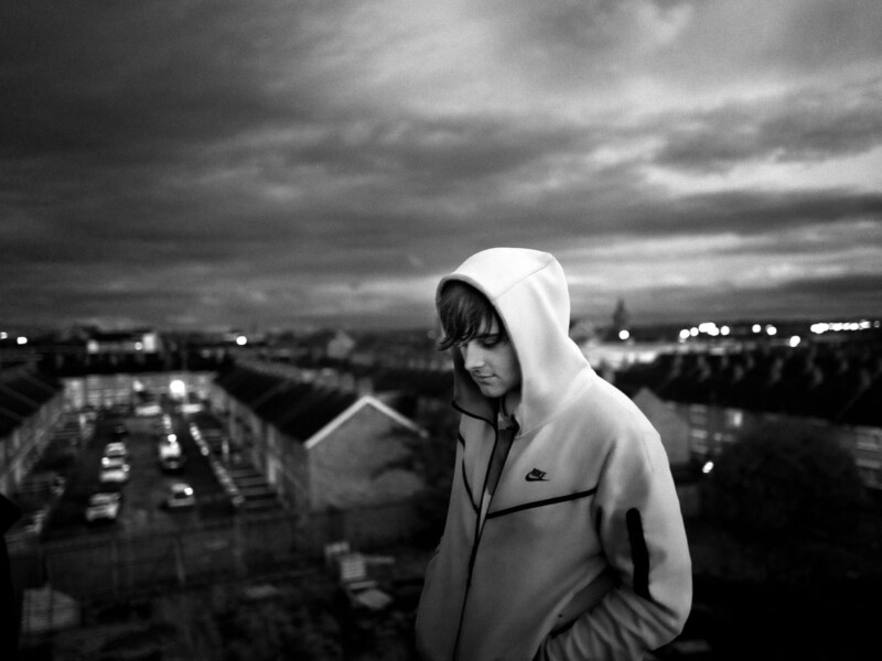 A young person wearing a hooded Nike jacket stands alone on a rooftop at dusk, looking down with hands in pockets. Rows of houses and parked cars stretch out in the dim, moody background.