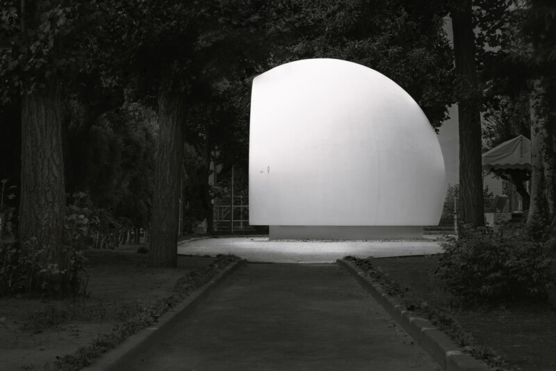 A large, white, minimalist half-sphere structure sits on a circular base in a park, surrounded by trees and shaded paths, with a tent visible in the background.