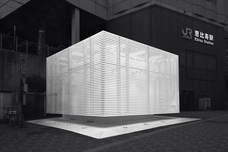 A modern, cube-shaped public restroom with illuminated horizontal slats stands outside Ebisu Station in Tokyo. The structure contrasts sharply against the dark building behind it.