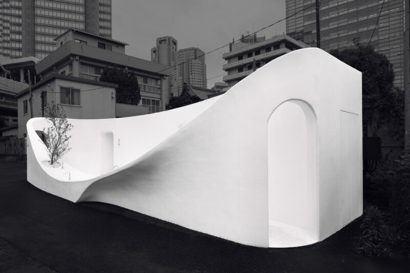 A modern, minimalist public restroom with smooth, curving white walls stands in an urban area surrounded by buildings and skyscrapers. The structure features arched doorways and a small tree planted inside.