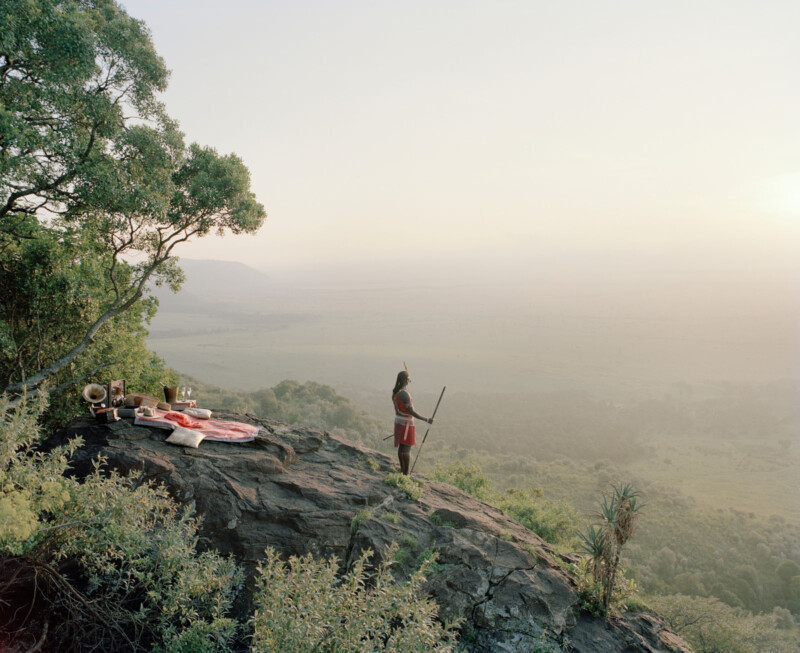 A person in traditional attire stands on a rocky cliff overlooking a vast valley. Nearby, a makeshift bed setup with blankets and cushions is arranged near the edge, surrounded by trees and greenery in soft morning light.