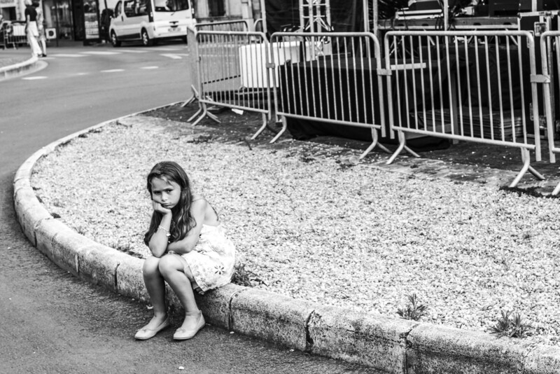 A young girl in a dress sits alone on a curb, resting her chin on her hand and looking unhappy. Metal barricades and parked vehicles are visible in the background. The photo is black and white.