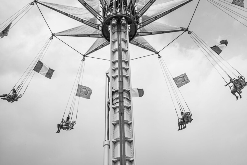 Black and white photo of a tall swing ride at an amusement park, with people sitting in swings suspended by long cables, and various flags attached above them, set against a cloudy sky.
