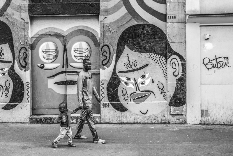 A man and a young child walk past a colorful mural featuring abstract human faces on a city wall, captured in black and white.