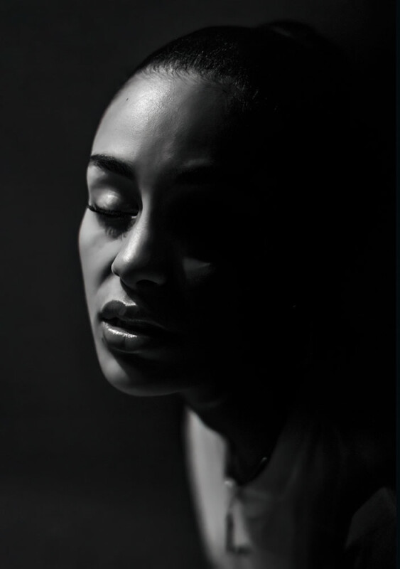 A woman with closed eyes is illuminated by soft, dramatic lighting from the side, creating deep shadows that partially obscure her face. The overall mood is serene and contemplative.