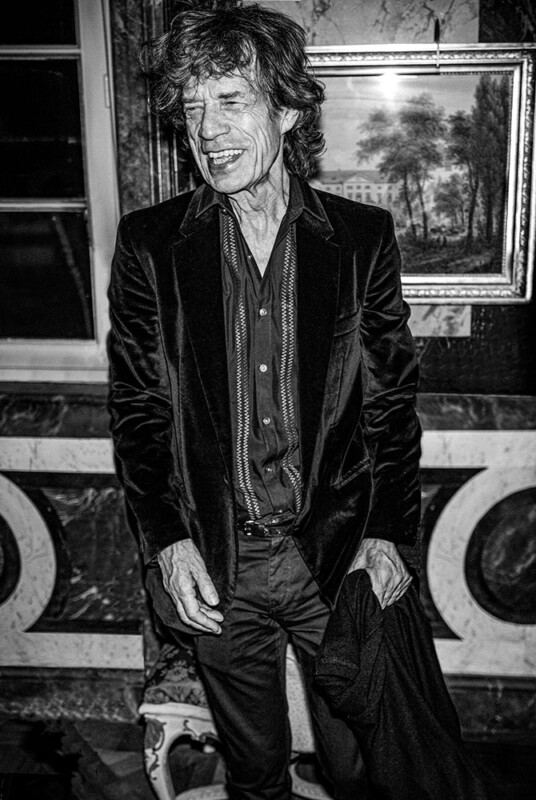 A man in a dark velvet suit stands indoors, smiling with eyes closed. The background features a framed landscape painting and ornate wall decor. The photo is in black and white.