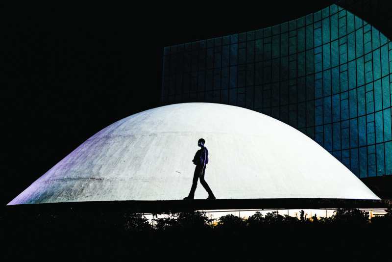 A person walks in front of a large, white dome-shaped structure at night, with a modern glass building in the background and dramatic contrast between light and shadow.