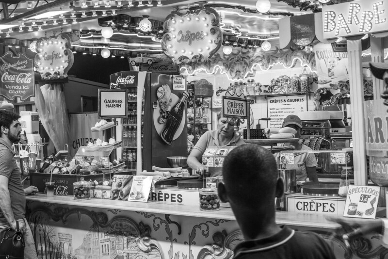 A busy, brightly decorated crepe stand at a fair, with signs in French, colorful lights, and people ordering and waiting in front. The scene is lively, with various treats and menu boards visible. The image is in black and white.