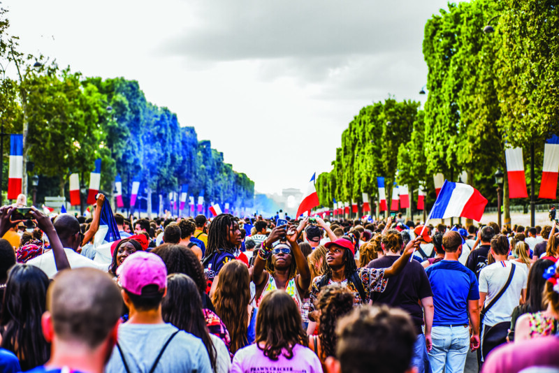 A large crowd gathers outdoors on a tree-lined street decorated with French flags, many people are taking photos and waving flags, with the Arc de Triomphe visible in the distance under a cloudy sky.