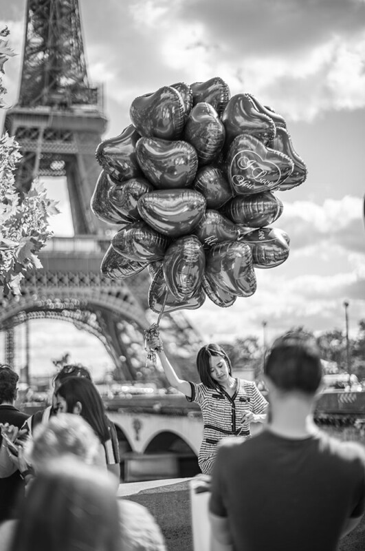 A woman holds a large bunch of heart-shaped balloons near the Eiffel Tower, surrounded by people. The cityscape and the iconic tower are in the background. The image is in black and white.