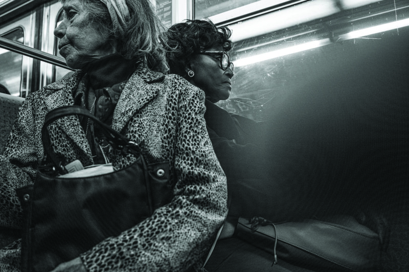 Two women sit side by side on public transit, facing different directions. One wears a leopard-print coat and headband, holding a handbag; the other wears glasses and dark clothing. The image is in black and white.