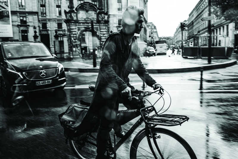 A person rides a bicycle on a wet city street, with raindrops visible on the camera lens. Cars and historic buildings appear in the background, creating a dynamic, urban scene in black and white.