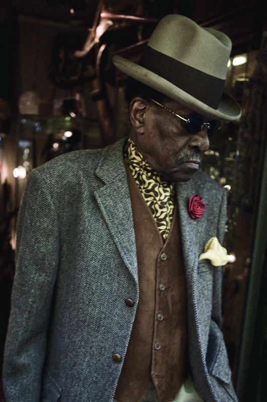 An older man wearing a grey herringbone blazer, brown vest, patterned scarf, sunglasses, and a fedora hat with a wide brim, accented with a red rose and yellow pocket square, stands in a dimly lit, stylish setting.