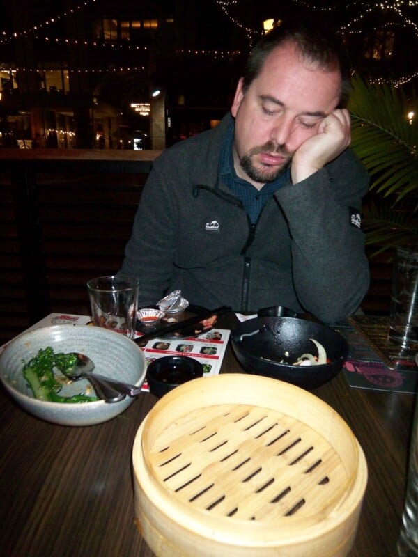 A man with a beard, wearing a dark jacket, is sitting at a table with various dishes. He appears sleepy, resting his head on his hand. The background has string lights and a plant is visible beside him.