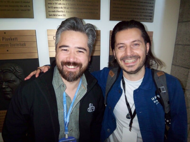 Two smiling men standing close together, one with gray hair and beard in a black jacket, the other with longer hair in a blue jacket. They are in front of a wall with metal plaques. Both have lanyards around their necks.