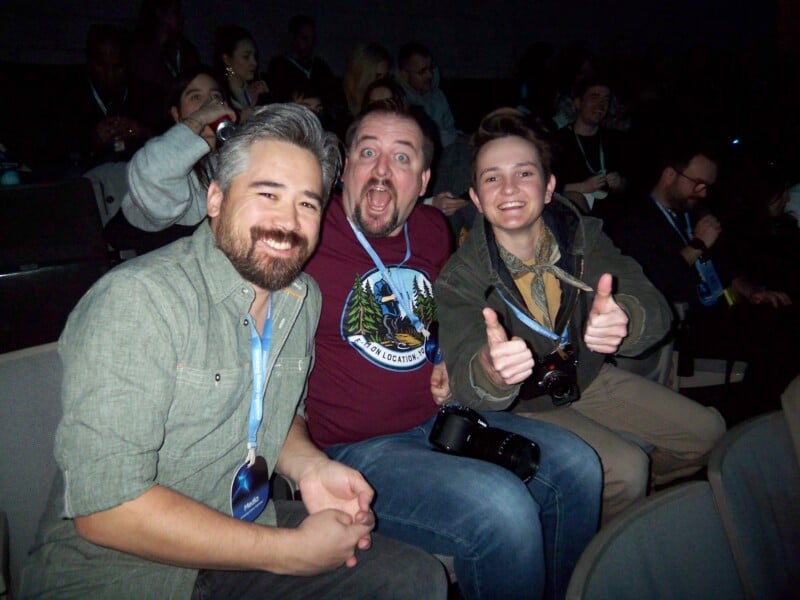 Three people are sitting in a theater, smiling at the camera. The person on the left has gray hair and a beard, the middle person is making a surprised face, and the person on the right gives two thumbs up. They all wear lanyards.
