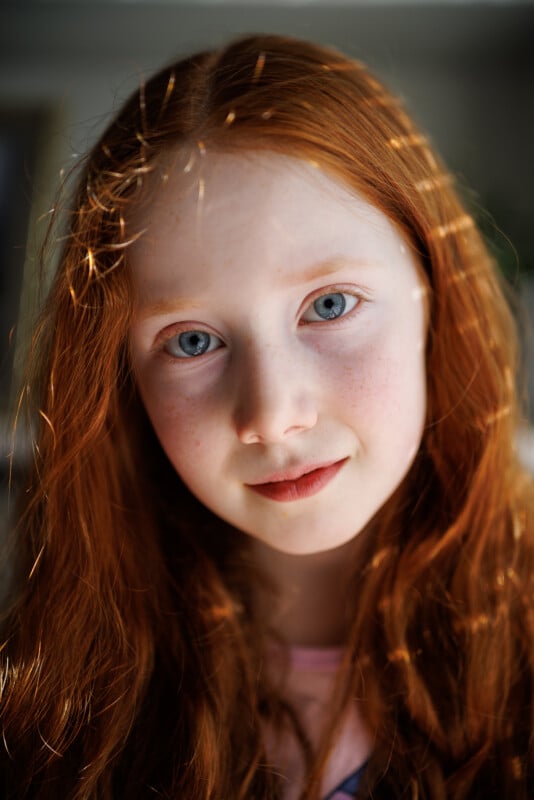 A young girl with long red hair and blue eyes looks directly at the camera. Sunlight catches her hair, creating a glowing effect. She has a gentle expression, and she's wearing a light pink top. The background is softly blurred.