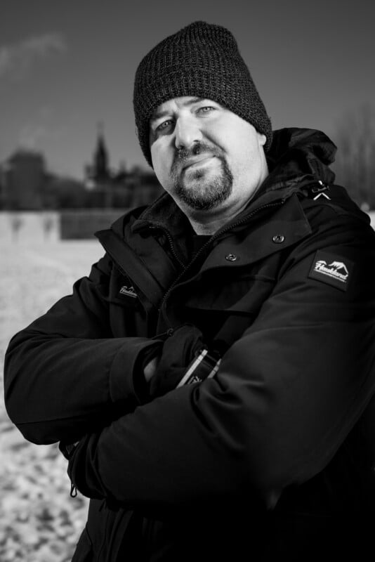 A man wearing a dark beanie and a coat stands outdoors with arms crossed. The background is a snowy landscape with buildings visible. The image is in black and white.