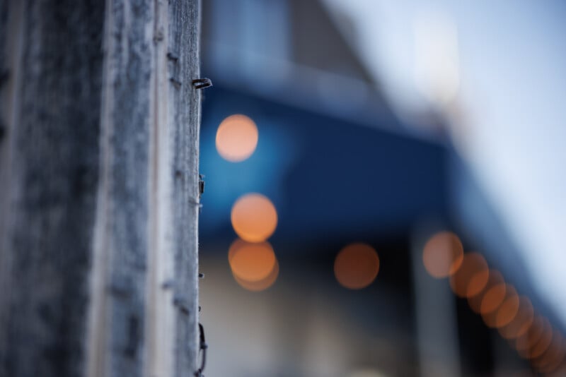 Close-up of a weathered wooden post with nails sticking out. In the background, circular orange lights are blurred against a blue and gray backdrop, creating a bokeh effect. The scene captures a mix of textures and soft light.