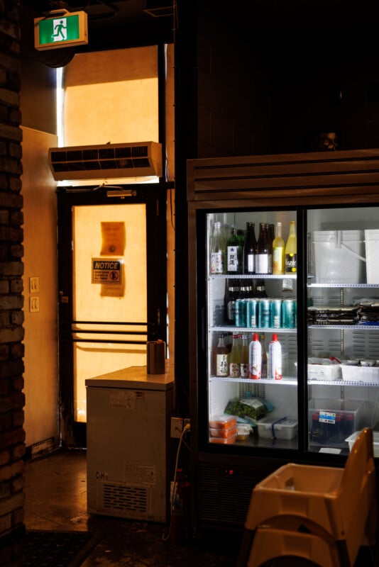 Interior of a dimly lit room with a glass refrigerator displaying drinks and food items. An exit door with a glowing green sign is partially open, letting in sunlight. A high chair and a small freezer are nearby.
