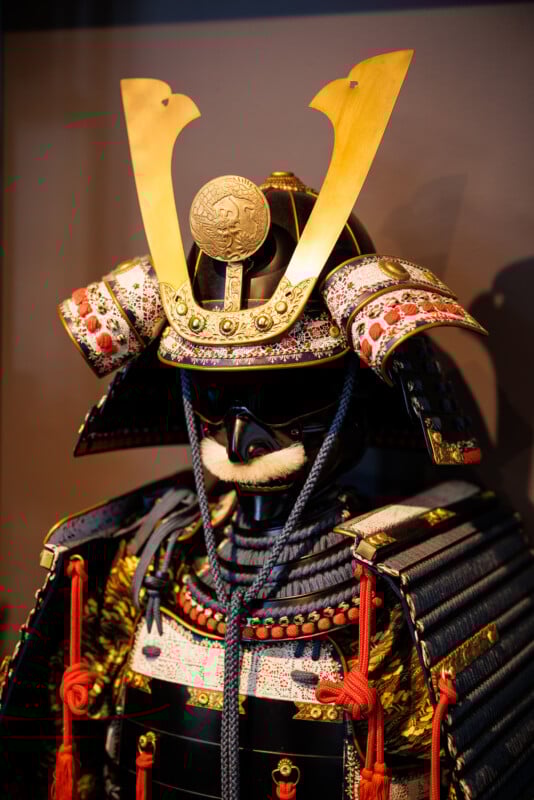 A traditional Japanese samurai armor set featuring an ornate helmet with golden antlers and intricate details. The armor is primarily black with red and gold accents, and includes a mask with a mustache.