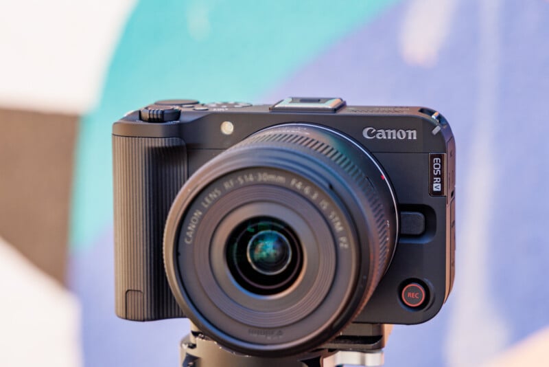 A Canon EOS R camera mounted on a tripod, focusing straight ahead. The lens details are visible, and the camera is set against a blurred background of various colors, including teal and black.