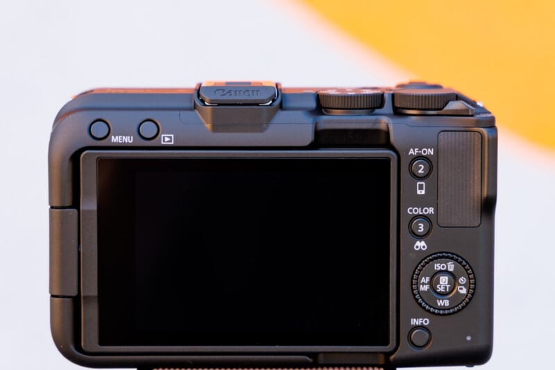 Rear view of a Canon camera showing the LCD screen, control buttons, and dials. The screen is off, and the buttons include AF-ON, MENU, and INFO. The camera is positioned against a blurred background with a gradient of white and orange.